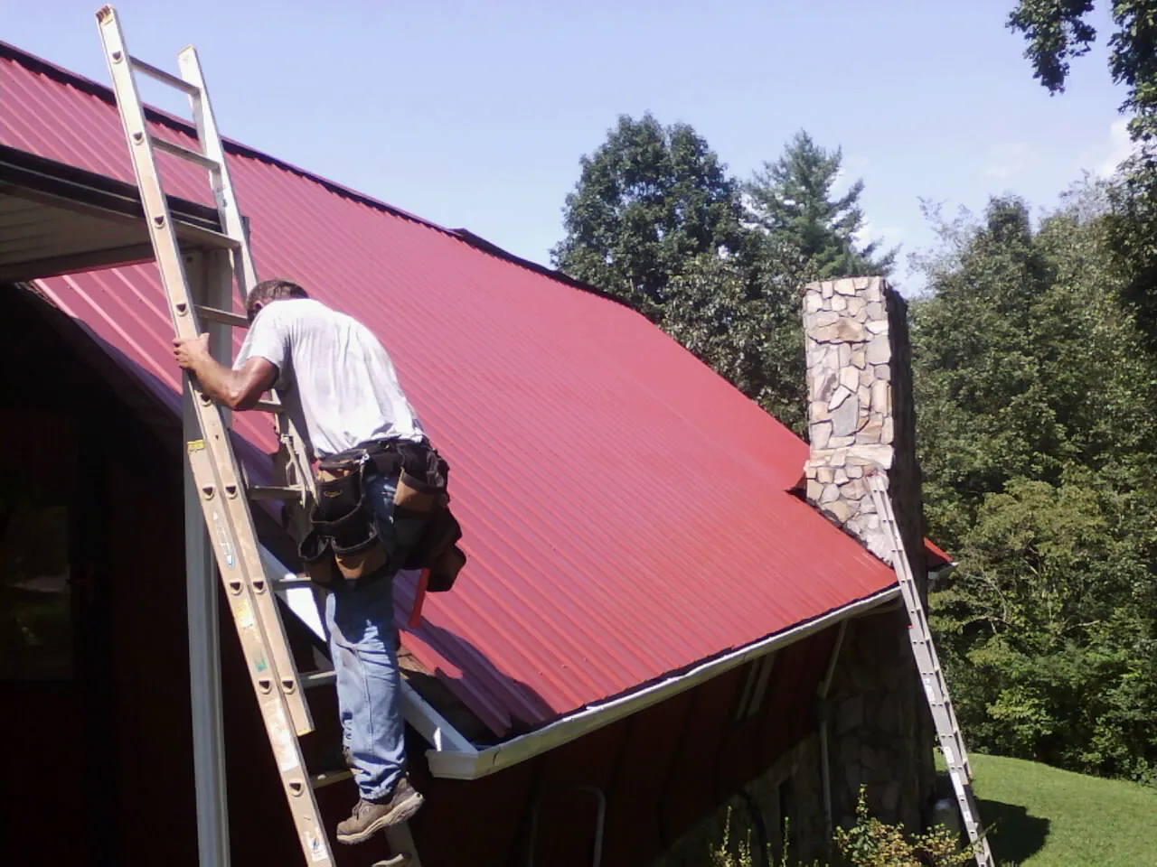 Roofer on a ladder beside a red metal roof and stone chimney — Soffit & Fascia Repair by Roofing Brothers in Burlington